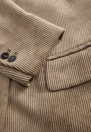 Smėlio spalvos riestinis audinys su siauru korduro tekstūra, dvi juodos sagos ir tvarkingai siūtas kišenė kairėje pusėje.