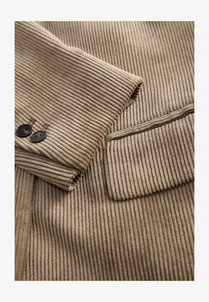 Smėlio spalvos riestinis audinys su siauru korduro tekstūra, dvi juodos sagos ir tvarkingai siūtas kišenė kairėje pusėje.