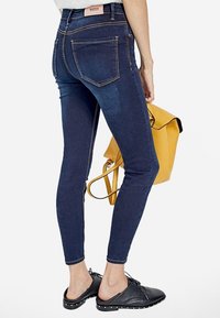 Jeans oscuros de cintura alta con bolsillos delanteros y traseros, costuras en contraste y corte ajustado, combinados con una mochila amarilla y zapatos negros.