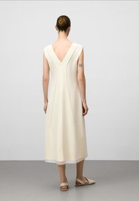 Cremfarbendes, ärmelloses Kleid mit einem V-Ausschnitt im Rücken, knielang, mit vertikalen Nähten und einem Reißverschluss auf der Rückseite. Glatte Textur.