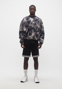 Νέος άνδρας που φοράει μαύρο και λευκό γραφικό φούτερ, μαύρο αθλητικό σορτς με λευκές ρίγες, λευκές κάλτσες και λευκά αθλητικά παπούτσια.