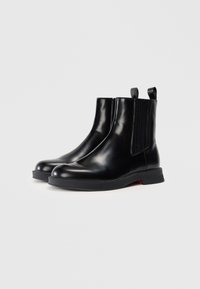Bottines en cuir noir avec panneaux latéraux élastiques, bout rond, languettes de tirage et semelle en caoutchouc légèrement texturée. Design épuré, détails minimaux.