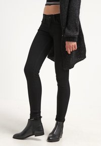 Jeans skinny negros con una cintura alta, con una textura de denim suave y costuras sutiles. Combinados con botas negras hasta el tobillo.