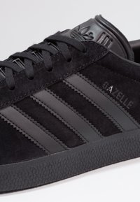 Svart mocka Adidas Gazelle sneakers med svarta läderremsor, texturerad sula och svarta snörningar.