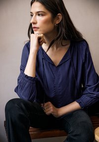 Top blu navy con scollo a V, maniche arricciate e pattina frontale. Il modello indossa jeans scuri, seduto su una superficie di legno, con la mano sul mento.