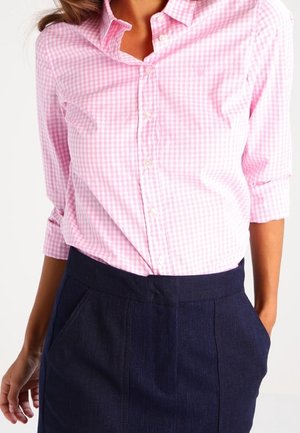 Kvinde iført en pink og hvid ternet skjorte med knapper og trekvartærmer, der er stoppet ind i et højtaljet navyblåt skørt med lommer.