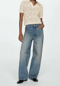 Polo en tricot beige à manches courtes avec motifs en losange, associé à un jean en denim bleu clair taille haute à jambes larges. Chaussures noires.