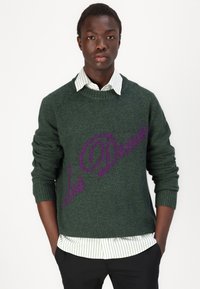 Dunkelgrüner Strickpullover mit gerippten Bündchen und Saum, der mit einem auffälligen lila Logodesign auf der Vorderseite gestaltet ist und über einem gestreiften Hemd getragen wird.