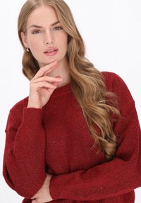 Rode gebreide trui met lange mouwen, ronde hals en een gestructureerde afwerking. Het model heeft lang golvend haar en steunt zachtjes haar hand op haar kin.