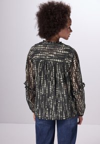 Transparente schwarze Bluse mit cremefarbenem Punktmuster, Rüschensäumen und gerafftem Rückendetail. Kombiniert mit blauen Jeans.