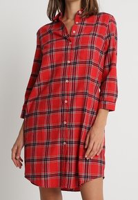 Robe chemise en flanelle rouge à motif carreauté, avec un devant boutonnière, un col et des manches trois-quarts. Texture douce, coupe ample.