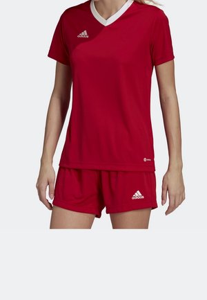 Vrouw draagt een rood Adidas sportshirt met witte V-hals en bijpassende rode korte broek, beide met Adidas-logo's.