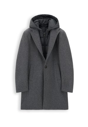 Homme portant un pardessus gris, une écharpe grise à motifs, un pantalon gris foncé, des lunettes de soleil noires et des baskets noires, debout devant un fond blanc.