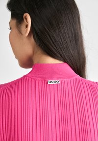 Åtsittande rosa ribbad tröja med hög hals. Har en liten svart "HUGO"-etikett på ryggen och visar upp en strukturerad tyg och design.
