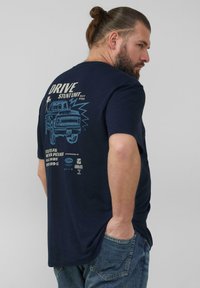 Granatowy t-shirt z krótkim rękawem z graficznym wzorem przedstawiającym pojazd i tekst na plecach, wykonany z bawełny, prezentowany na osobie.