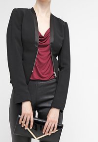 Blazer negro con ribete de satén, con cierre de cremallera, combinado con una parte superior drapeada burdeos y pantalones de cuero negros. Sosteniendo un bolso de mano negro.