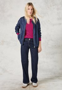 Jeans in denim scuro, maglione bordeaux e una leggera giacca bomber blu navy con polsini a costine. Sneakers bianche completano l'outfit.