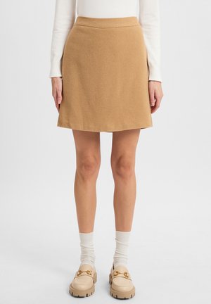 Ein hellbraßer A-Linien-Rock aus weichem Stoff, mit glatter Textur und einem Saum, der bis zur Mitte des Oberschenkels reicht. Kombiniert mit beigen Loafers und weißen Socken.