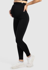 Leggings de maternité noirs, fabriqués à partir d'un tissu extensible ; design taille haute avec une texture lisse. Portés avec des baskets blanches.