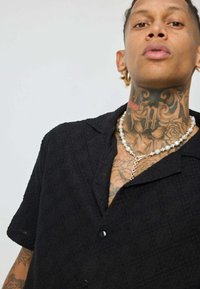 Chemise noire à manches courtes avec un motif texturé, comprenant un col et des boutons noirs. Le modèle porte un collier de perles et a des tatouages visibles.