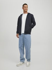 Marinefarbener Zip-Hoodie mit Kapuze und Kordelzug, kombiniert mit hellblauen, locker sitzenden Jeans und weißen Sneakers mit grauen Akzenten.