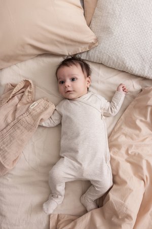 Lys beige baby onesie på en myk, nøytral sengebakgrunn. Omgivelsestekstiler inkluderer et krøllete teppe og glatte puter med tekstur.