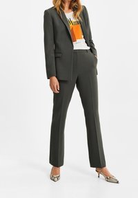 Tmavě zelený oblek na míru se sakem na jedno zapnutí s klopami s výřezem a rovnými kalhotami. Kombinováno s bílým tričkem s potiskem a botami na podpatku s hadím vzorem.