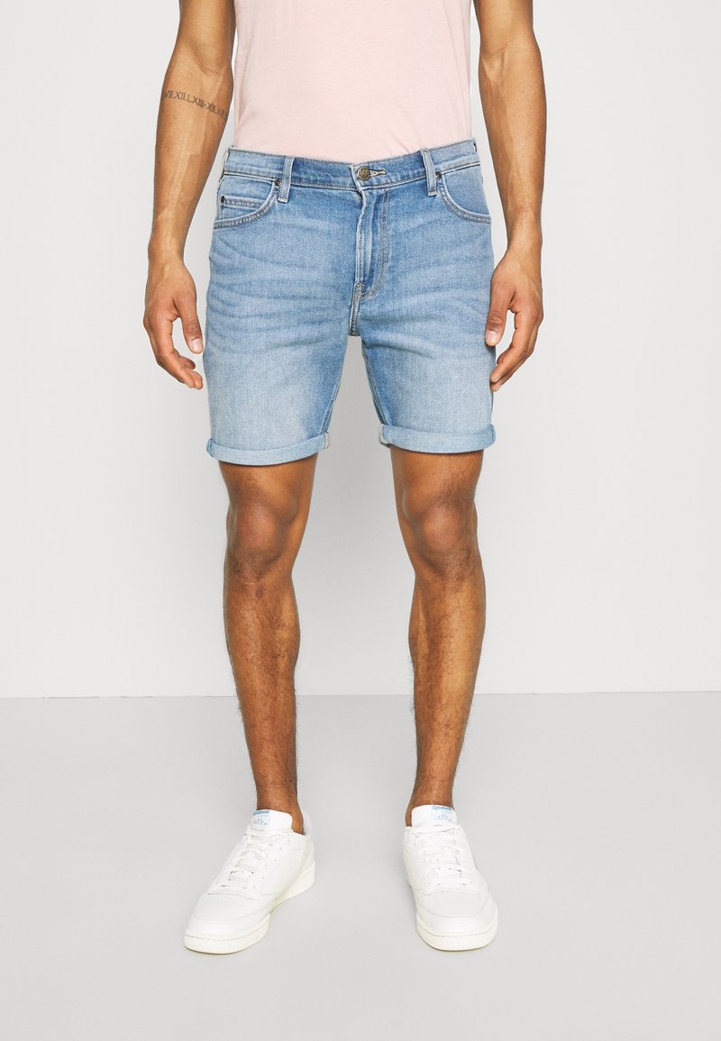 Homme portant un short en jean bleu clair retroussé, des baskets blanches et une chemise rose pâle, debout devant un fond uni.