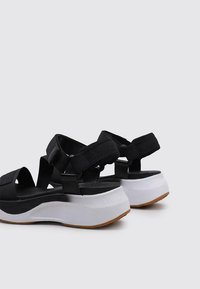 Sandalias negras con múltiples correas ajustables, una plantilla negra acolchada y una suela media blanca, que cuenta con una suela exterior marrón texturizada.