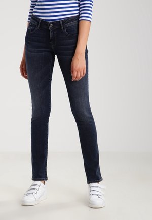 Femme portant un jean skinny foncé, des baskets blanches à scratch et une chemise à manches longues rayée bleu et blanc, debout.
