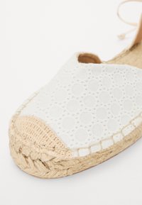 Weiße Espadrilles mit kreisförmigen Mustern auf dem Stoffobermaterial, Jutesohle und beigem Zehenkappen. Das Design zeichnet sich durch eine gewebte Textur und leichte Schnürung aus.