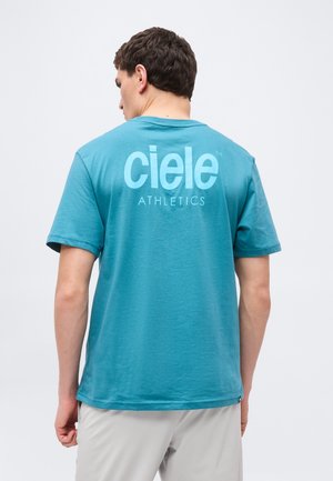 Muž v tyrkysovom tričku s krátkym rukávom s veľkými písmenami "ciele ATHLETICS" na chrbte, oblečený v svetlo sivých nohaviciach, zobrazený zozadu.