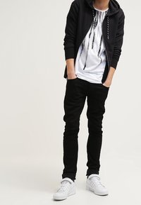 Schwarze Zip-Hoodie, weißes Grafik-T-Shirt mit schwarzem Design, schwarze Jeans und weiße Sneaker. Vor einem hellen, neutralen Hintergrund.
