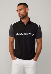 Camisa polo negra con logotipo blanco "HACKETT", acentos rojos en el cuello y mangas cortas grises. Hecha de una tela suave, combinada con pantalones blancos.