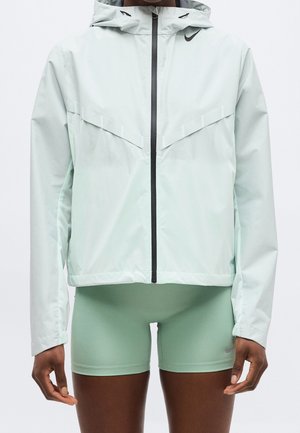 Personne portant une veste à capuche zippée vert clair et un short ajusté assorti, debout les bras détendus le long du corps.