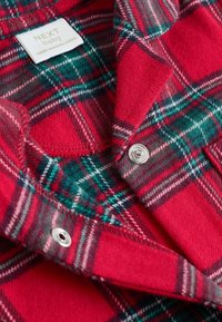 Chemise en flanelle rouge avec un motif à carreaux verts et noirs, boutons-pression argentés et étiquette tissée. Texture douce avec un col plié.