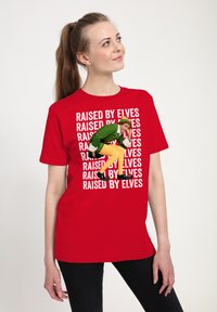Červené bavlněné tričko s bílým nápisem "VYCHOVÁVÁNI ELFY" opakovaně potisknutým, s postavou v zeleném na přední straně, která má na sobě žluté kalhoty.