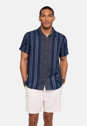 Hombre con camisa de manga corta azul marino con rayas verticales y pantalones cortos blancos, de pie con las manos en los bolsillos.