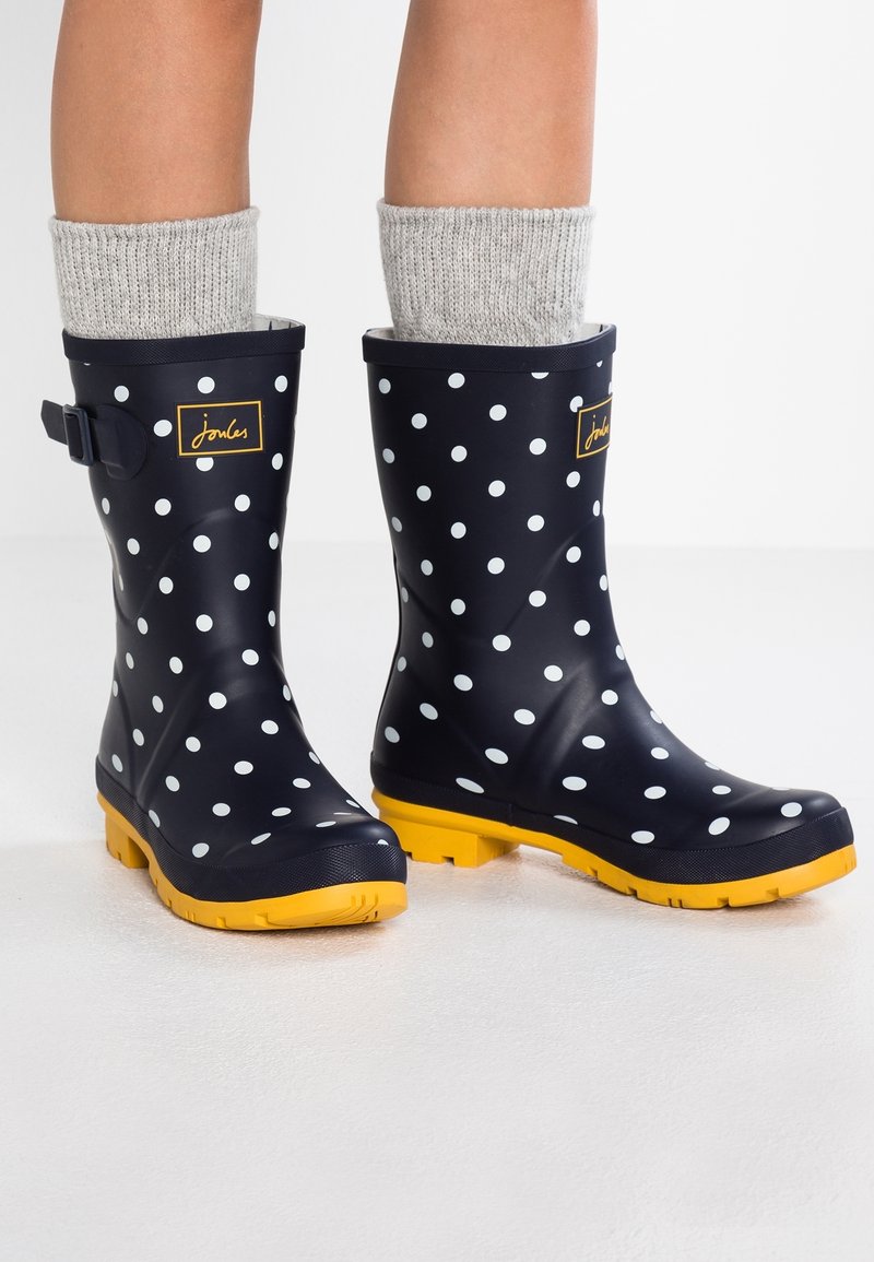 Stivali da pioggia in gomma blu navy con pois bianchi, suole gialle e una calza lavorata grigia che spunta dalla parte superiore. Dettaglio con cinturino regolabile al lato.