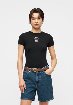 Giovane che indossa una maglietta nera aderente con piccoli loghi Cadillac e Tommy Hilfiger abbinata a pantaloncini di jeans a vita alta e cintura marrone.