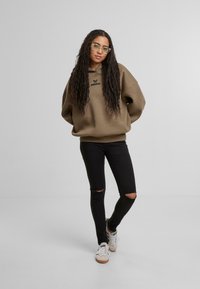 Oversized olívazöld kapucnis pulóver logóval, fekete, szakadt skinny farmernadrággal és fehér sportcipőkkel párosítva. A modellen áttetsző szemüveg van.
