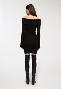Abito nero a coste con spalle scoperte, maniche lunghe, silhouette aderente e lunghezza sopra il ginocchio. Abbinato a stivali neri alti fino alla coscia.