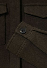 Detail tmavě hnědého vlněného kabátu s knoflíkovými manžetami, bočními kapsami a měkkým, strukturovaným povrchem. Klasický design s dokonanými detaily.