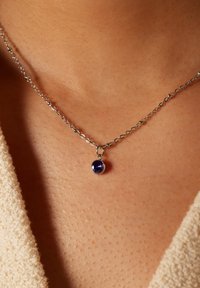 Zilveren ketting met een ronde blauwe edelsteen hanger, rustend tegen een gestructureerde huidoppervlak. De ketting is delicaat en verfijnd.