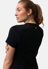 Black short-sleeve top with a smooth texture, round neckline, and a logo detail on the back. Model shown from the side, hair tied back.