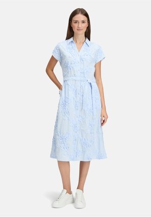 Femme debout, main dans la poche, portant une robe midi bleu clair brodée de fleurs et des baskets blanches sur fond blanc.