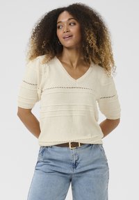 Maglione corto a maniche corte color crema con scollatura a V, strisce testurizzate e dettagli ad occhiello. Abbinato a jeans chiari in denim e una cintura marrone.