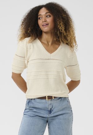Krem pulover s kratkimi rokavi in V-izrezom, textured stripe in očesni detajli. V kombinaciji z svetlo jeanskimi hlačami in rjavim pasom.