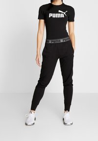 Kobieta ubrana w czarną koszulkę Puma, czarne spodnie dresowe Puma z charakterystycznym paskiem oraz białe sneakersy Puma, stojąca z rękami w kieszeniach.