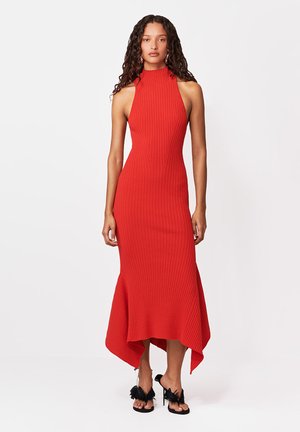 Kobieta o długich kręconych włosach, ubrana w czerwoną, bezrękawnikową sukienkę prążkowaną z asymetrycznym dołem oraz czarne sandały na obcasie z piórowymi detalami.